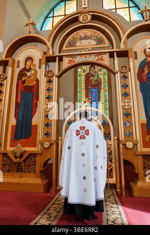 Un prêtre en vêtements ornés dirige un service religieux devant un autel magnifiquement décoré avec des icônes et des vitraux dans un c orthodoxe Banque D'Images