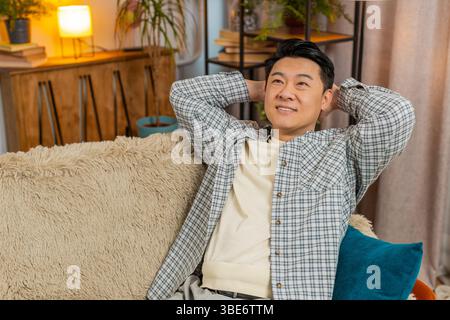Heureux homme chinois d'âge moyen insouciant assis confortablement avec la main derrière la tête sur le canapé dans le salon à la maison. Souriant mec coréen se relaxant sur le canapé pensant regarder autour dans la chambre de l'appartement. Banque D'Images