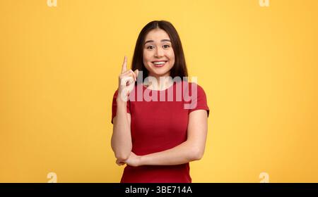 Femme souriante en chemise rouge pointant vers le haut contre un fond jaune vif Banque D'Images
