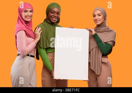 Trois musulmanes en hijab tenant une affiche vide, fond jaune Banque D'Images