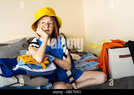 Jeune femme dans la tenue bleue et chapeau seau jaune se trouve entouré de vêtements se sentant submergé par la fatigue de décision et l'encombrement de placard essayant de le faire Banque D'Images
