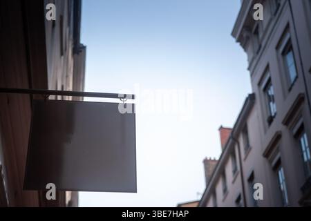 Un panneau carré blanc gris est suspendu à une façade sur une rue étroite, laissant un espace de copie pour l'image de marque sur le ciel doux et le bâtiment de style français floue Banque D'Images