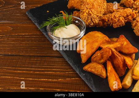 L'assiette comprend des morceaux de poulet frits croustillants ainsi que des quartiers de pommes de terre dorées et un petit bol de trempette crémeuse, sur une table en bois foncé. Banque D'Images