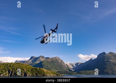 Hélicoptère en vol au-dessus d'une vallée glaciaire et de pentes montagneuses abruptes en Norvège, avec un ciel bleu clair et un paysage de fjords expansif. Banque D'Images
