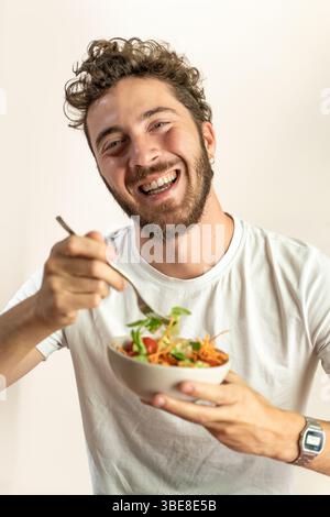 Jeune homme appréciant la salade saine à la maison souriant avec fraîcheur et énergie Banque D'Images