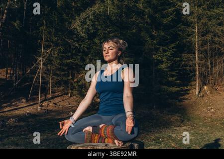 Une femme pratiquant le yoga effectue une séance assise en position lotus sur une souche d'arbre, méditant seule dans des vêtements décontractés lors d'une journée d'automne ensoleillée dans le parc. Bonjour Banque D'Images