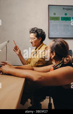 Deux collègues femmes discutant des détails du projet à un bureau. L'atmosphère est professionnelle, collaborative et enthousiaste, démontrant le travail d'équipe et Banque D'Images