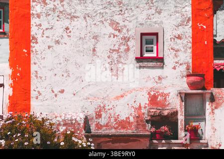 Charmante façade rustique d'une ancienne maison méditerranéenne avec des murs texturés blancs et rouges altérés par les intempéries, de petites fenêtres, des pots en argile et des fleurs en fleurs sous Banque D'Images