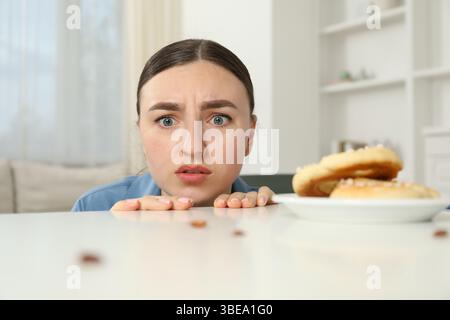 Jeune femme choquée regardant des cafards et des bretzels sur une table blanche à l'intérieur. Problème de ravageur Banque D'Images
