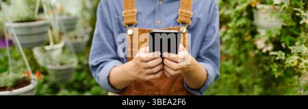 Une jeune jardinière nourrit ses plantes dans une serre colorée, vérifiant son téléphone pour des conseils, bannière Banque D'Images