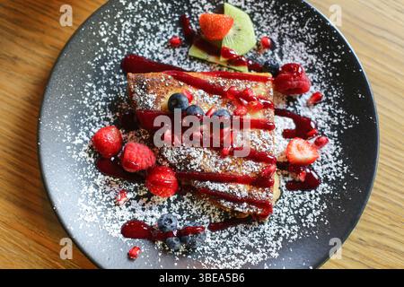 Petit déjeuner sucré avec des baies. Crêpes au sucre en poudre. Présentation élégante des aliments. Garniture de fruits sur assiette foncée. Concept de dessert matinal. Banque D'Images