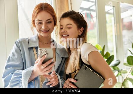 Deux jeunes femmes élégantes rient et apprécient la compagnie des autres tout en vérifiant le téléphone. Banque D'Images