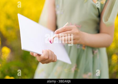 Gros plan de la peinture d'enfant fille sur papier blanc vierge avec de la peinture rose en utilisant un bâton en bois debout dans le champ de fleurs jaunes, créant vibrant en plein air Banque D'Images