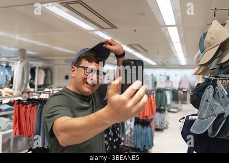 Un homme prend un selfie avec son smartphone, essayant un nouveau chapeau dans un magasin de vêtements. Consommateur masculin, achats de seconde main, appel vidéo avec des amis. Banque D'Images