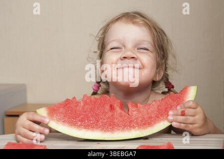 Petite fille de 2 ans, assise à la table de la cuisine et mange avec plaisir la pastèque. Visages putride Banque D'Images