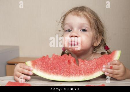 Petite fille de 2 ans, assise à la table de la cuisine et mange avec plaisir la pastèque. Visages putride Banque D'Images
