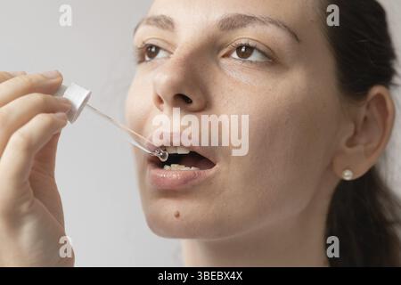 Portrait de jeune femme aux cheveux foncés prenant un supplément d'huile avec compte-gouttes en verre. Huile de médecine de chanvre CBD appliquée sur la langue. Banque D'Images