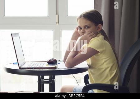 Une jeune fille bouleversée est assise à une table à la maison, fait des devoirs et regarde avec mécontentement Banque D'Images