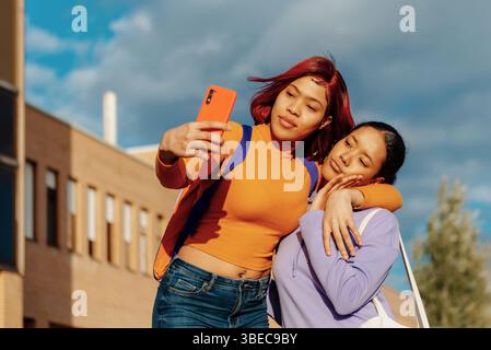 Deux jeunes femmes latines, l'une avec les cheveux roux et l'autre avec les cheveux noirs, sont vues profiter d'une journée ensoleillée sur un campus universitaire tout en prenant un selfie avec un s. Banque D'Images