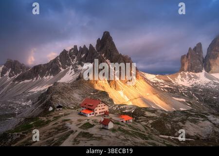 Vue aérienne des montagnes, maison et église au coucher du soleil en automne. Vue de dessus de hautes roches, neige, ciel violet avec nuages. Les sommets montagneux en automne. Tre Cime Banque D'Images