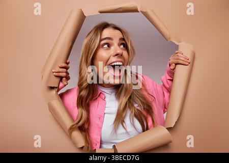 Une jeune femme joyeuse aux cheveux longs et fluides jetant un regard ludique à travers un mur de papier déchiré, incarnant parfaitement l'excitation et la surprise tout en étant habillée Banque D'Images