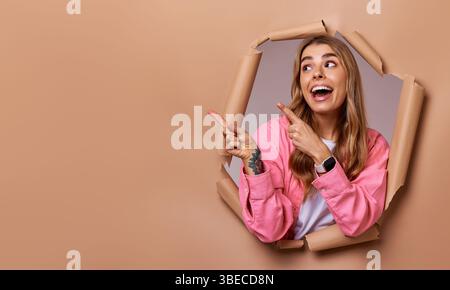 Une jeune femme joyeuse aux cheveux blonds éclatants souligne joyeusement tout en émergeant d'un fond de papier déchiré de manière créative, mettant en valeur son éne sans bornes Banque D'Images