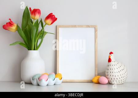 Décorations de maison de Pâques sur table. Maquette de cadre photo, poulet décoratif, oeufs de Pâques, vase de tulipes sur fond de mur blanc. Banque D'Images
