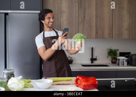 Chef prenant des photos de brocoli avec téléphone dans la cuisine, entouré de légumes frais. Banque D'Images