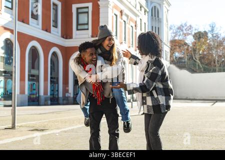 Trois amis joyeux multi-ethniques profitant d'une journée ensoleillée à l'extérieur, l'un donnant un tour de piggyback tandis que l'autre discute Banque D'Images