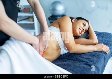 Masseuse professionnelle effectuant un massage relaxant sur l'abdomen du client souriant, favorisant le bien-être et les soins du corps dans l'environnement spa Banque D'Images
