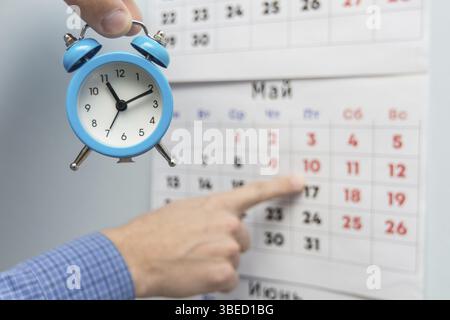 Une main tient un petit réveil, dans l'arrière-plan d'une part de points de longs week-ends et jours fériés sur un calendrier mural Banque D'Images