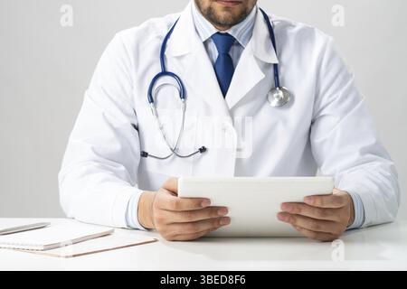 Gros plan des mains de médecin masculin à l'aide d'un ordinateur tablette. Médecin en uniforme médical blanc avec stéthoscope assis à son bureau. La technologie moderne en examina Banque D'Images