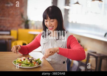 Femme asiatique assise à la table en bois dans le café, coupant la salade sur l'assiette avec une fourchette et un couteau Banque D'Images