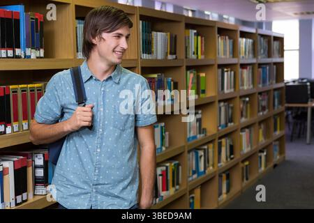 Étudiant masculin debout dans la bibliothèque portant un sac à dos à côté d'étagères en bois, espace de copie Banque D'Images