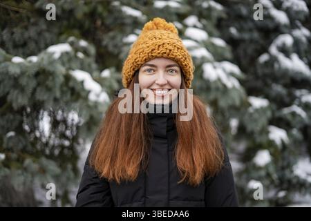 Femme souriante se situant dans les bois. Portrait de style de vie de jeune fille aux cheveux rouges souriante regardant la caméra. Concept de jour de Noël enneigé. Banque D'Images