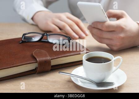 Homme en chemise blanche à l'aide d'un smartphone comme assis au bureau avec une tasse de café. Application professionnelle et technologie mobile. Navigation Internet et communication Banque D'Images