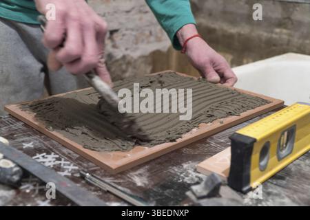 L'ouvrier applique de l'adhésif de ciment sur les carreaux. Travaux de finition, mise au point floue. La technologie de pose de carreaux Banque D'Images