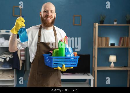 Sourire jeune homme barbu portant des gants et un tablier tenant panier avec des produits de nettoyage montrant un spray détergent. Femme de ménage masculine dans les tâches domestiques. Househ Banque D'Images