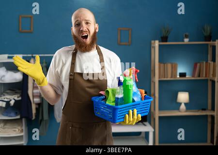 Bel homme barbu tenant des produits de nettoyage avec une expression surprise geste apologétique, disant désolé tout en se tenant debout dans le salon. Ménage, ho Banque D'Images