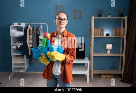 rire jeune femme confiante portant tablier et gants tenant des produits de nettoyage regardant loin avec expression naturelle debout dans le salon à Banque D'Images