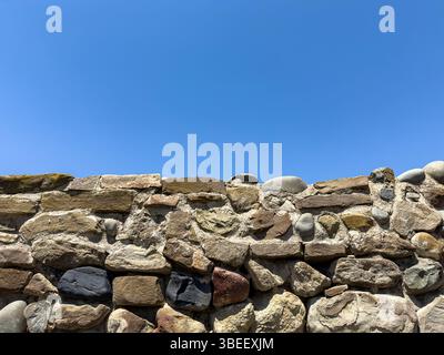 Vieux mur de pierre rustique avec des roches irrégulières et une texture rugueuse sous ciel bleu clair, lumière du jour, fond naturel pour la conception ou l'architecture. Personne. Banque D'Images