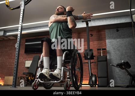 Individu avec des tatouages s'entraînant dans le gymnase, en utilisant des bandes de résistance tout en étant assis dans un fauteuil roulant. Équipement et dispositifs visibles en arrière-plan Banque D'Images