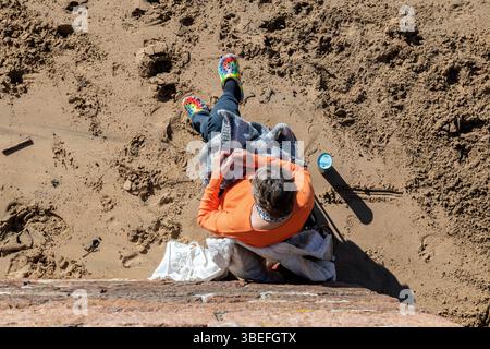 Une femme dans une chemise orange est assise sur la plage, tricotant avec des chaussures colorées, profitant d'une journée de détente au bord de la mer. Banque D'Images