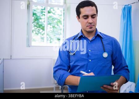 Médecin écrivant des notes sur le presse-papiers bleu portant le stéthoscope en clinique par le feuillage de fenêtre et le rideau Banque D'Images