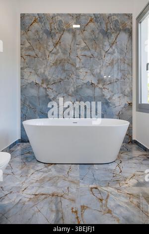 Luxueuse salle de bains moderne avec baignoire blanche sur pied placée contre un mur impressionnant de carreaux de marbre poli Banque D'Images