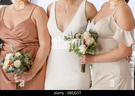 3 femmes élégamment habillées, probablement une mariée avec un décolleté en v profond habillée et ses demoiselles d'honneur en satin et beige clair, tenant de petits bouquets Banque D'Images