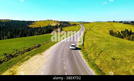 Voiture sur une route sinueuse à travers Green Hills. Vue aérienne d'une voiture blanche conduisant le long d'une route sinueuse qui traverse des collines verdoyantes et des forêts. Banque D'Images
