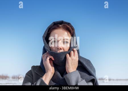 Une femme se tient près d'un lac gelé en hiver, enveloppée dans une écharpe grise, se protégeant du froid. Le ciel bleu vif est clair, mettant en évidence le Banque D'Images