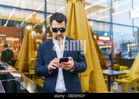 Homme d'affaires portant un costume et des lunettes de soleil, concentré sur son smartphone tout en se tenant à l'extérieur dans un cadre urbain moderne. Construction en verre brillant et réfléchissant Banque D'Images