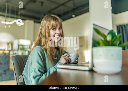 Une jeune fille blonde joyeuse appréciant son café dans un café confortable. Elle porte un pull vert clair, souriant chaleureusement tout en tenant une tasse, créant un a accueillant Banque D'Images